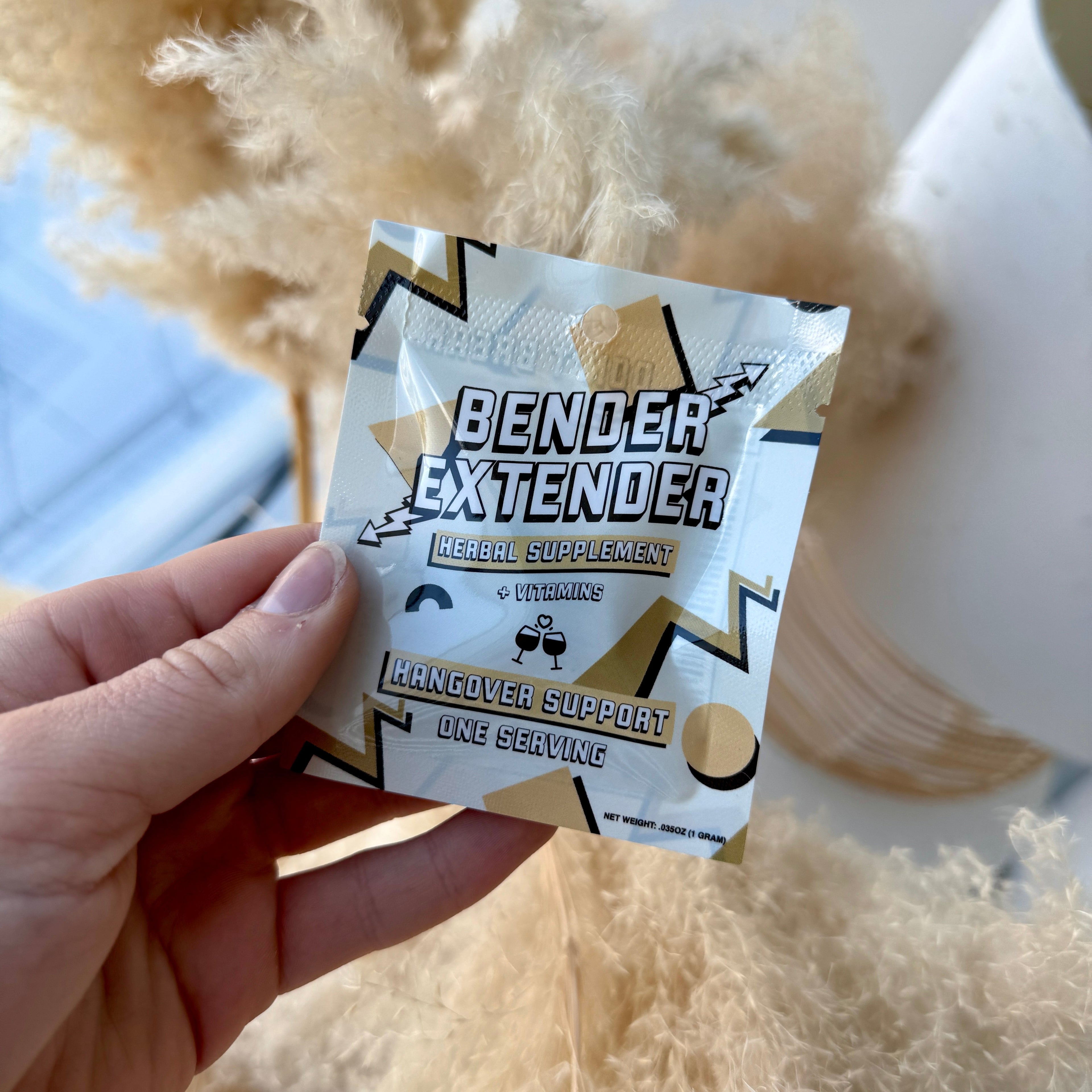Hand holding a 'Bender Extender' supplement packet with decorative dried plants in the background
